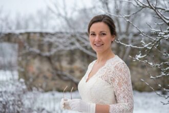 Mariée en robe ivoire tenant ses lunettes à monture dorée dans un jardin enneigé en hiver